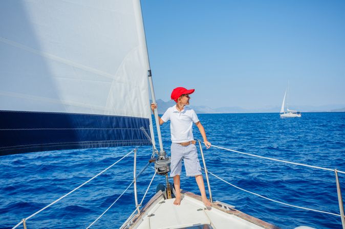 Junge mit roter Kappe steht vorne auf einem Segelboot und schaut auf das Meer