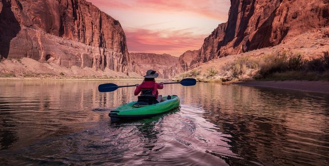 Eine Person sitzt in einem Grünen Kanu und schaut auf einen pinken Sonnenauf/untergang