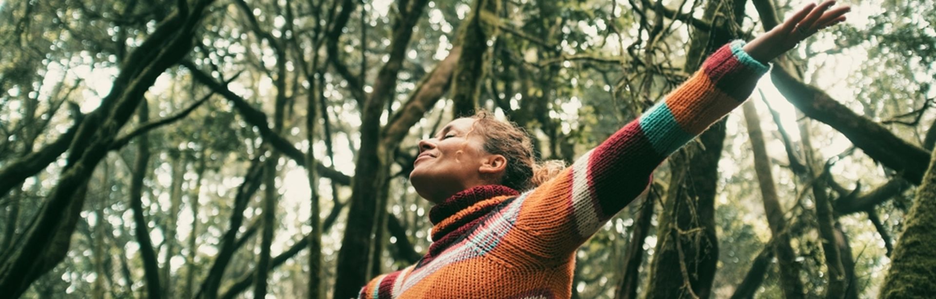 Eine Frau breitet glücklich die Arme aus und hat die Augen geschlossen, Sie steht in einem Wald voller dichter Bäume