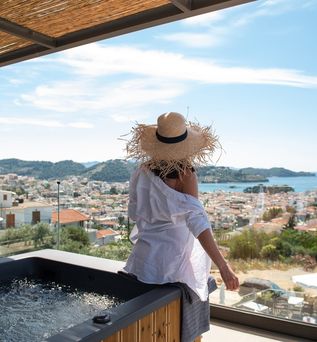Eine Frau mit Sonnenhut sitzt auf dem Rand eines Whirlpools auf einem Balkon und blickt runter Richtung der Stadt