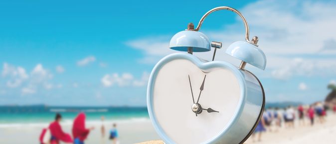 Auf einem Sandhügel steht ein hellblauer, herzförmiger Wecker, Ein Strand mit vielen Menschen ist verschwommen im Hintergrund zu sehen